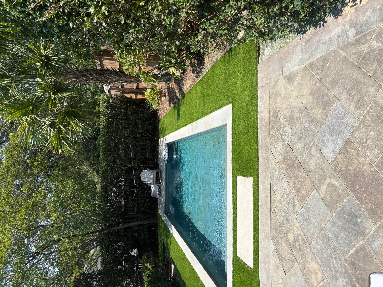 Synthetic turf pool deck with white trim, herringbone pavers, and mature landscaping